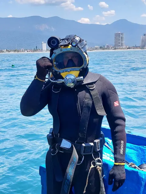 Commercial diver wearing a helmet and diving suit, with the coast in the background.