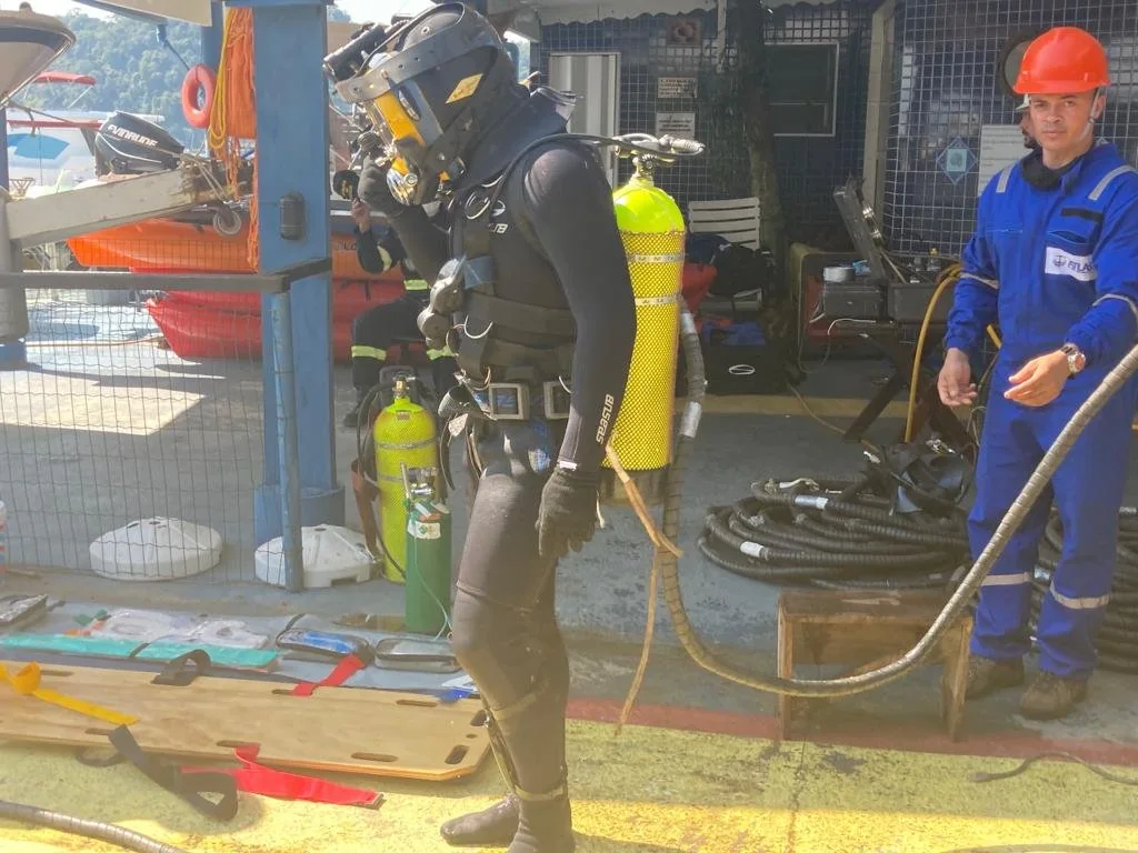 Mergulhador com capacete e equipe de apoio preparando os equipamentos para a operação.